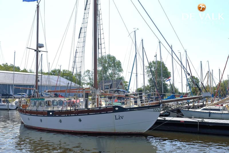 Een witte zeilboot met de naam "Liv" ligt afgemeerd in een haven, met andere boten en gebouwen op de achtergrond.