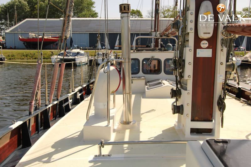 Een dek van een boot met een schoorsteen, een mast en een kajuit, met andere boten en gebouwen op de achtergrond.