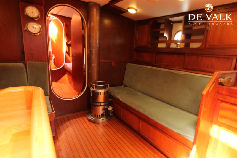 De foto toont een interieur van een boot met houten wanden en vloer, een groene bank, een kachel en een doorgang.