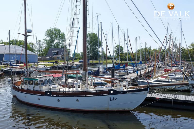 Een witte zeilboot met de naam "Liv" ligt afgemeerd in een jachthaven met vele andere boten en een gebouw op de achtergrond.