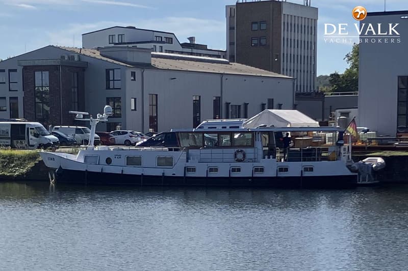 Een witte boot met een donkere romp ligt aangemeerd aan een kade met gebouwen en geparkeerde auto's op de achtergrond.