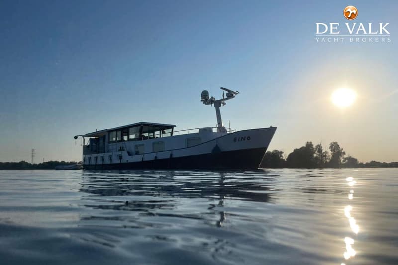 Een witte boot met de naam "GINO" ligt op het water, met de zon laag aan de horizon en een logo van "DE VALK YACHT BROKERS"…