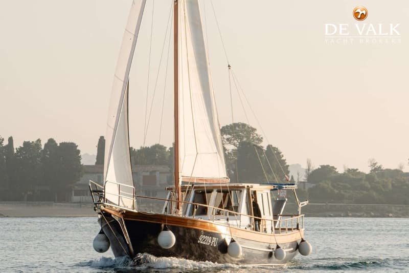 Een donkerblauwe zeilboot met witte zeilen en een houten opbouw vaart op het water, met een kustlijn op de achtergrond.