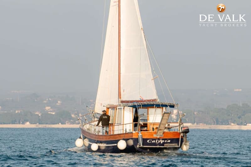 Een zeilboot met de naam "Calypso" vaart op open water, met een persoon aan boord. — Custom Built Houseboat luxe motor