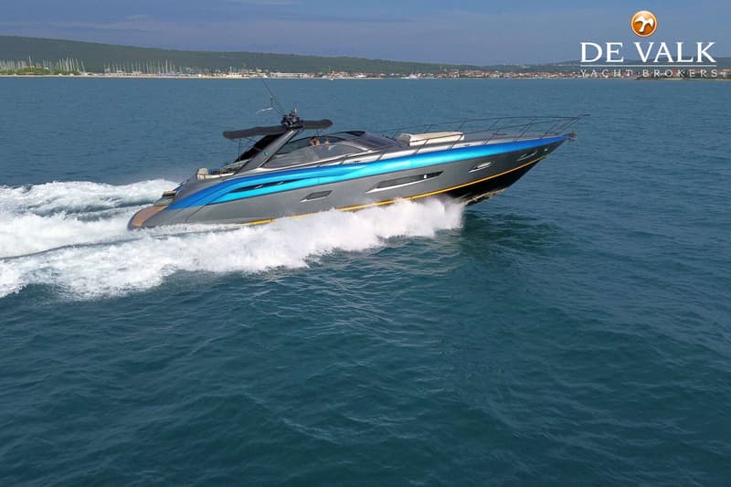 Een grijze en blauwe motorboot vaart snel over het water, met een kustlijn en bebouwing op de achtergrond.