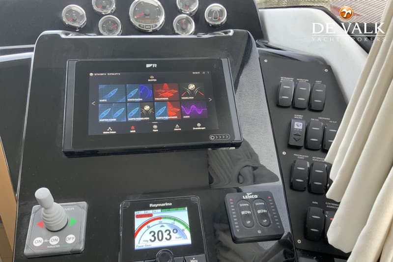 De foto toont een deel van een dashboard met diverse meters, een groot digitaal scherm, een joystick en schakelaars.