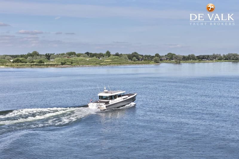 Een grijze motorboot met witte opbouw vaart op een waterweg, met een groene oever en blauwe lucht op de achtergrond.