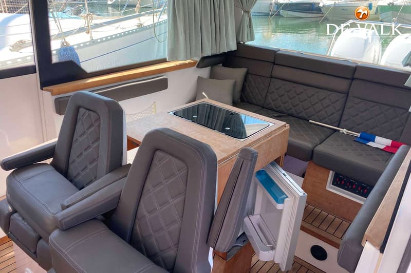 Het interieur van een boot met twee grijze stoelen, een houten tafel met kookplaat, een bank en een open koelkast.