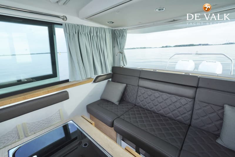 De foto toont het interieur van een boot met een donkergrijze bank, ramen met gordijnen en een kookplaat. — Axopar 37 AC