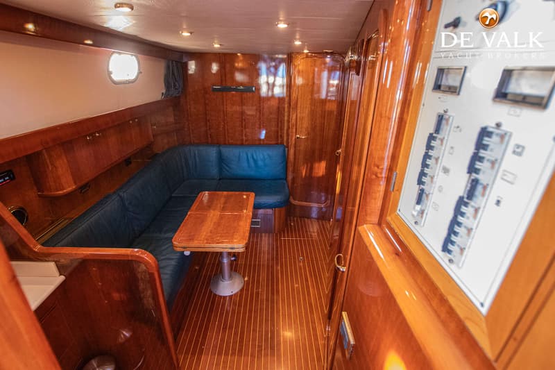 Een interieur van een boot met een L-vormige blauwe bank, een houten tafel en een schakelpaneel aan de rechterkant.