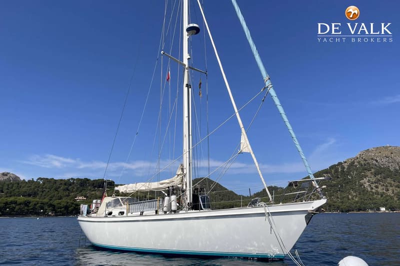 Een witte zeilboot met een blauwe streep op de romp ligt voor anker in een baai met beboste heuvels op de achtergrond.