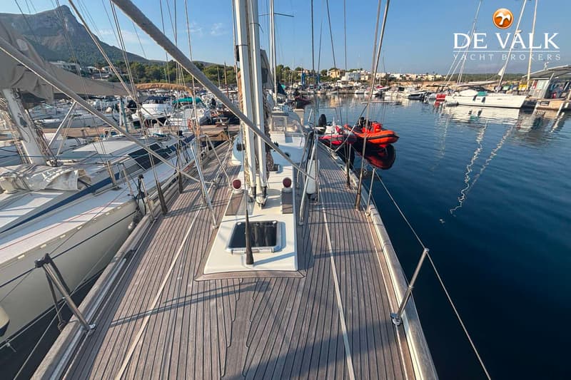 Een houten dek van een zeilboot met een open luik, omringd door andere boten in een haven met bergen op de achtergrond.