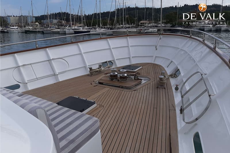 Een houten dek met twee lieren en bolders, een wit schip met gestreepte kussens en een haven op de achtergrond.