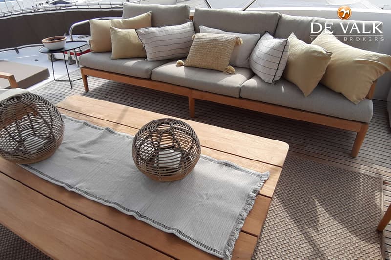 Een houten tafel met een grijze tafelloper en twee rieten lantaarns staat voor een bank met kussens op een teakhouten dek.