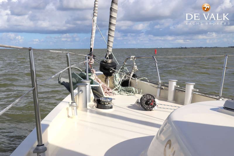 De foto toont het voordek van een zeilboot met touwen, lieren en een rolfokinstallatie, varend op water onder een bewolkte…