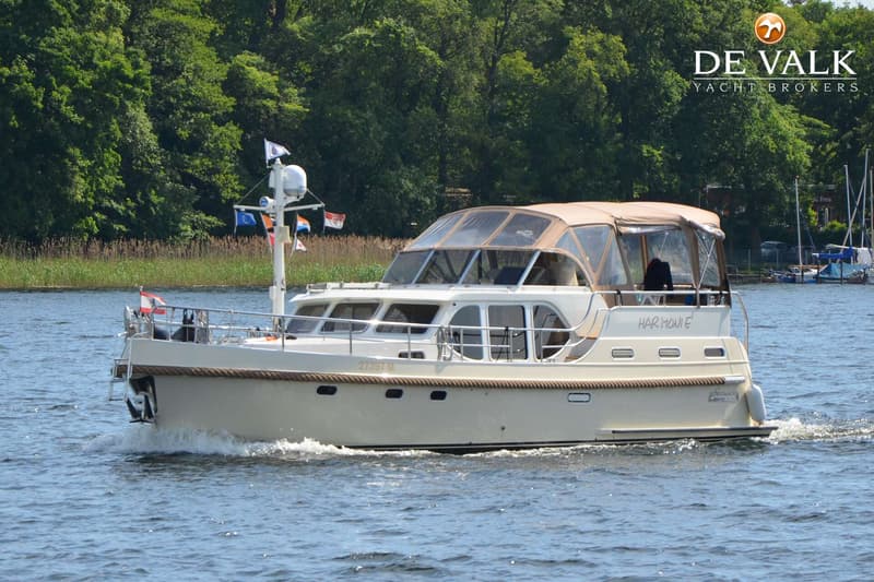 Een beige motorjacht met een bruine kap vaart op het water, met bomen en riet op de achtergrond.
