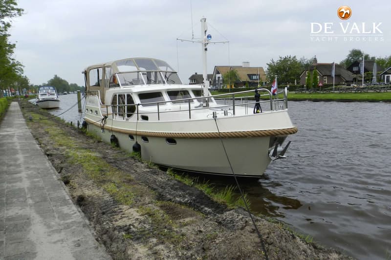 Een beige motorjacht ligt afgemeerd langs een waterweg met huizen op de achtergrond.