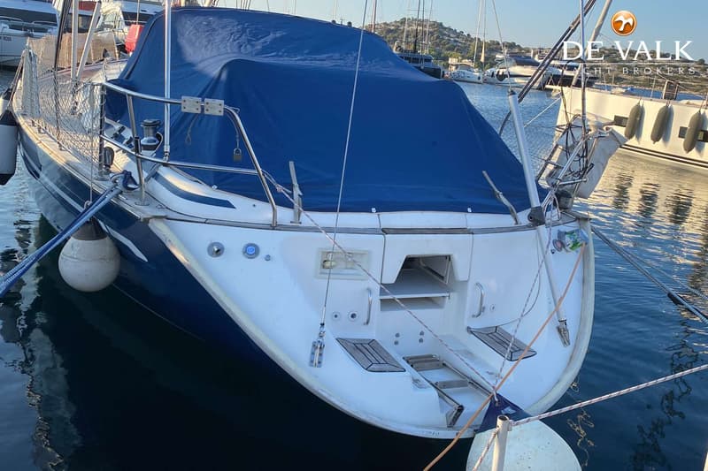 Een blauw-witte zeilboot met een blauw dekzeil ligt afgemeerd in een haven. — Bavaria 47 Cruiser