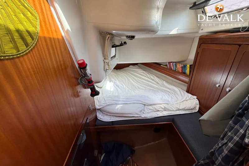 Een kajuit met een bed met wit dekbed, houten wanden en kasten, en een boekenplank. — Bavaria 47 Cruiser