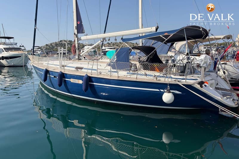 Een blauwe zeilboot met de naam "UNIT" is afgemeerd in een haven, omringd door andere boten. — Bavaria 47 Cruiser
