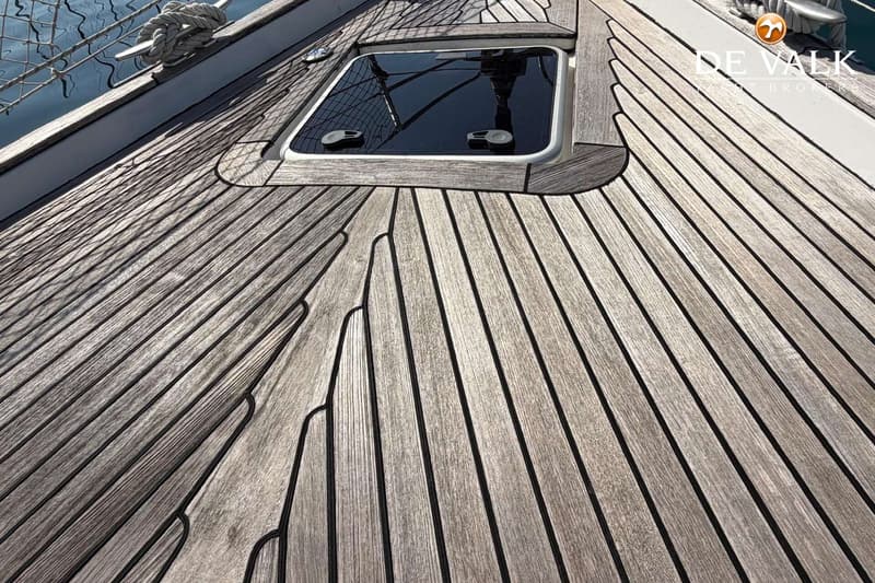 Een teakdek met een luik in het midden, een kikker met touw links en een "De Valk Yacht Brokers" logo rechtsboven.