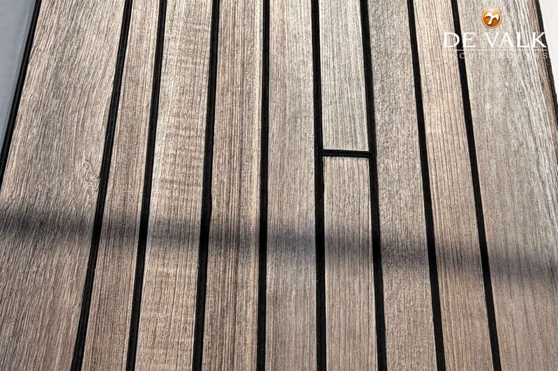 De foto toont een close-up van een houten dek met zwarte naden en het logo van "DE VALK YACHT BROKERS". — Bavaria 47 Cruiser