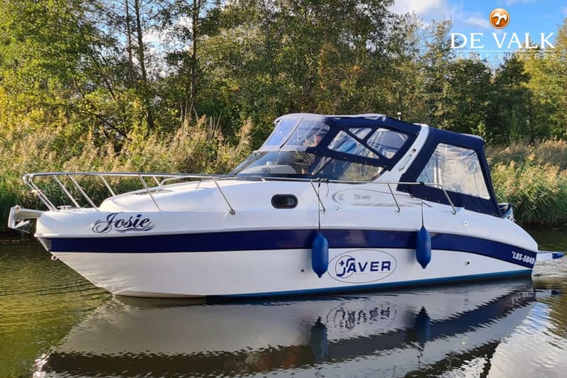Een witte motorboot met blauwe accenten en de naam "Josie" op de boeg, drijvend op water met bomen op de achtergrond.