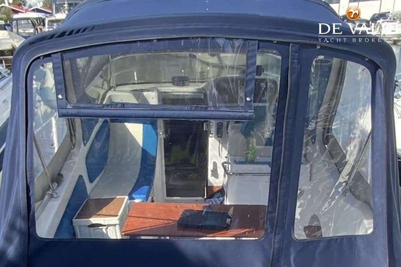 Een blauwe bootkap met doorzichtige ramen toont een interieur met een houten tafel en blauw-witte kussens.