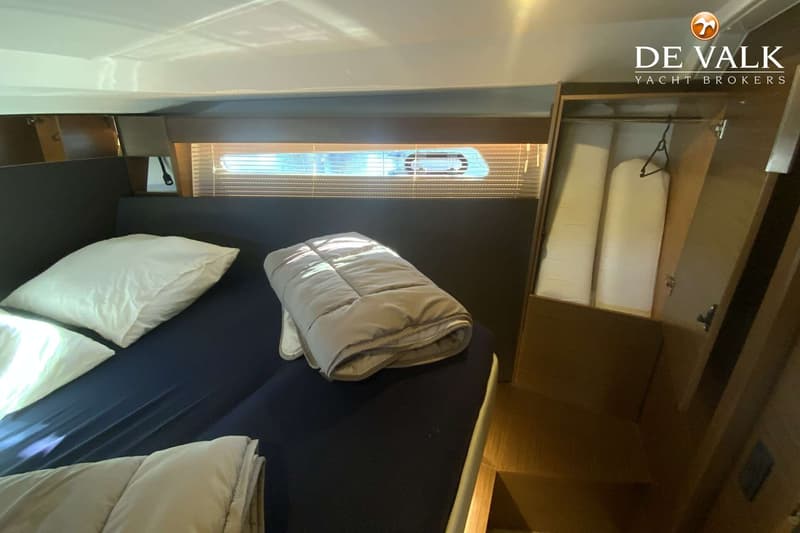 Een kajuit met een bed, een raam met jaloezieën en een open kast met kledinghangers en opgerolde matrassen.