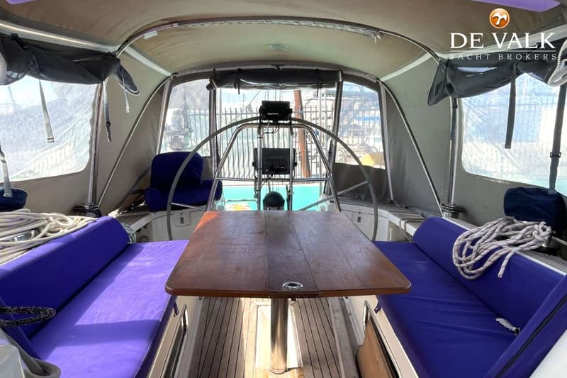 De foto toont de kuip van een zeilboot met een houten tafel, paarse kussens en een stuurwiel met navigatie-instrumenten.