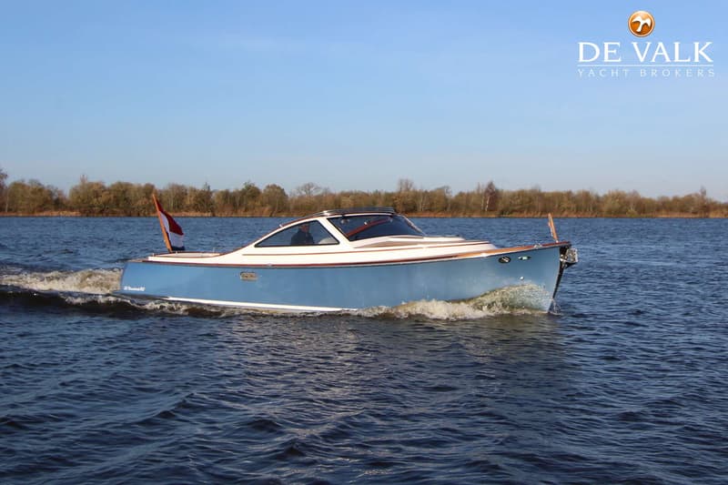 Een blauw-witte motorboot vaart op een waterweg met een bosrijke oever op de achtergrond onder een heldere blauwe lucht.