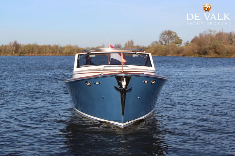 Een blauw-witte motorboot met een Nederlandse vlag vaart op een waterweg met bomen op de achtergrond.