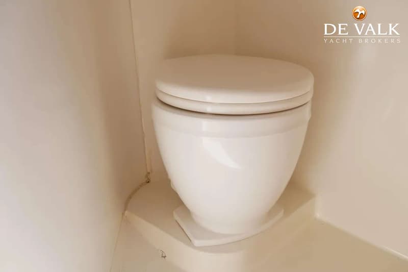 Een witte scheepstoilet met gesloten deksel staat in een hoek van een lichte ruimte.