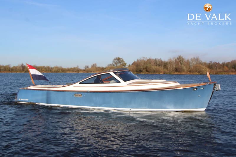 Een blauw-witte motorboot met een Nederlandse vlag vaart op een meer met bomen op de achtergrond. — Long Island 33 Runabout