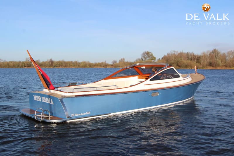 Een blauw-witte boot met houten accenten vaart op een waterweg met bomen op de achtergrond. — Long Island 33 Runabout