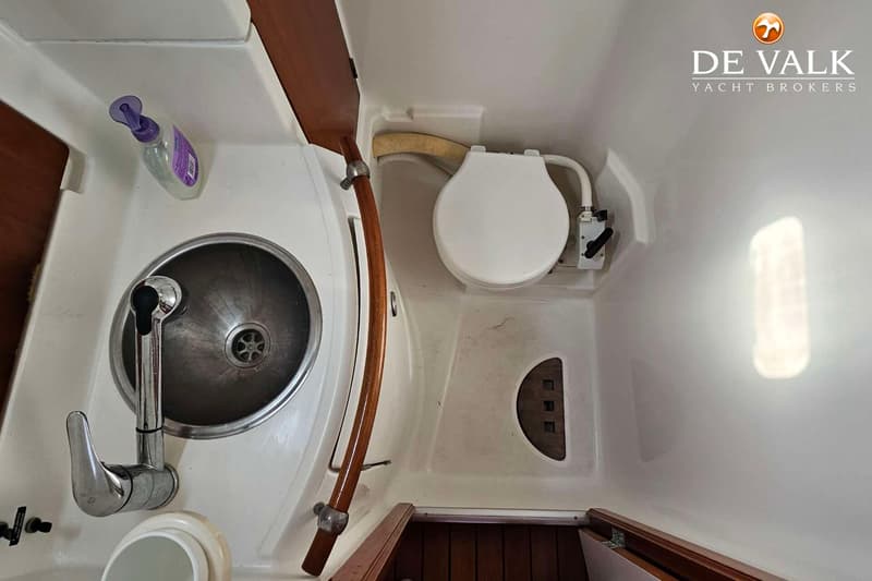 Een boottoiletruimte met een wasbak, kraan, zeepflesje en een toilet met een houten handgreep ernaast.