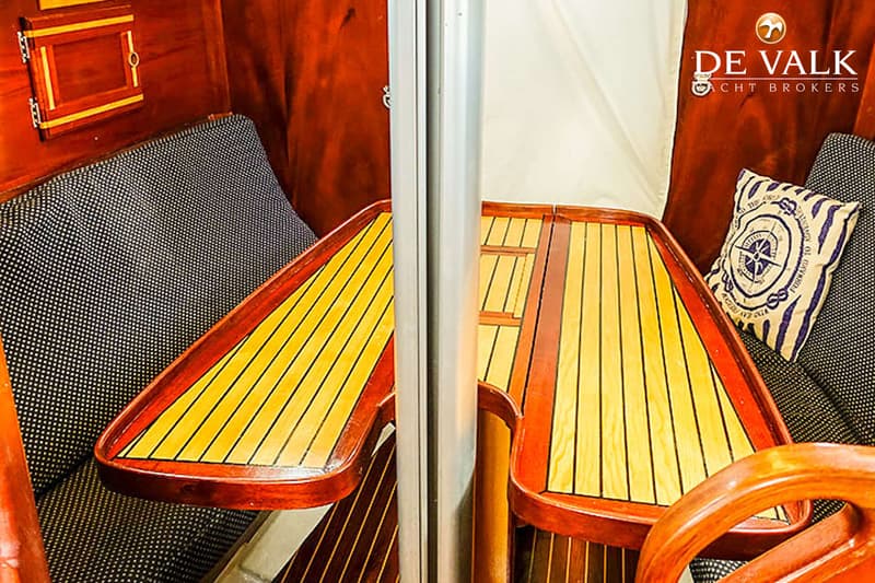 Een interieur van een boot met houten wanden, een tafel met twee uitklapbare bladen en donkere banken met kussens.