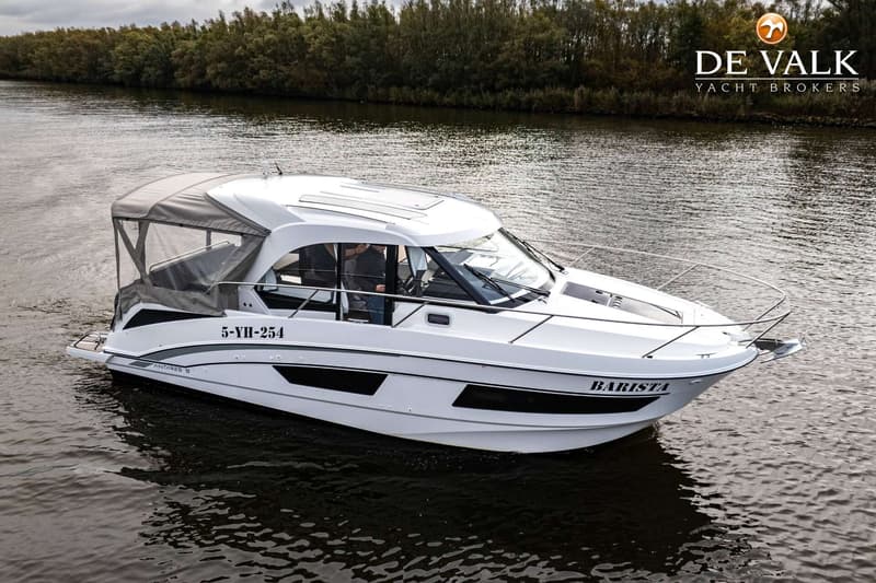 Een witte motorboot met de naam "BARISTA" en registratienummer "5-YH-254" vaart op water met een beboste oever op de…