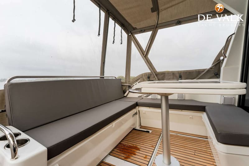 Een bootinterieur met een lange bank met grijze kussens, een tafel en een houten vloer.