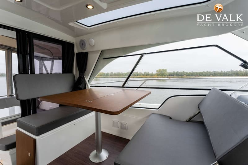 De foto toont een interieur van een boot met een houten tafel, grijze zitbanken en ramen die uitkijken op water en land.