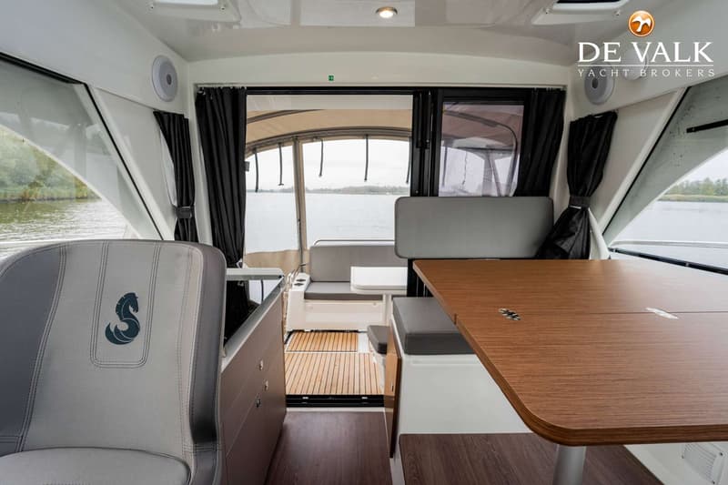 De foto toont het interieur van een boot met een tafel, zitbanken, een stuurstoel en uitzicht op water.