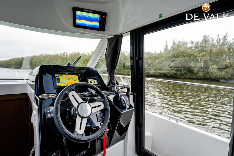 Een stuurhut van een boot met een stuur, navigatieschermen, een gashendel en uitzicht op water en bomen.