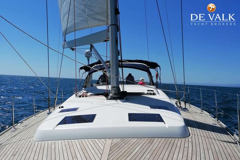 Een zeilboot met een houten dek en een wit dek, met een mast en zeilen, vaart op een blauwe zee onder een heldere hemel.