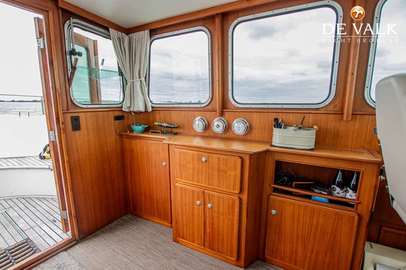 De foto toont het interieur van een boot met houten lambrisering, ramen, een open deur naar buiten en diverse opbergkasten.