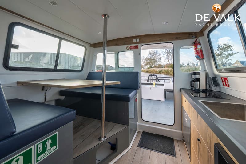 De foto toont het interieur van een boot met een zithoek, een tafel, een keukenblok en een open deur naar het achterdek.