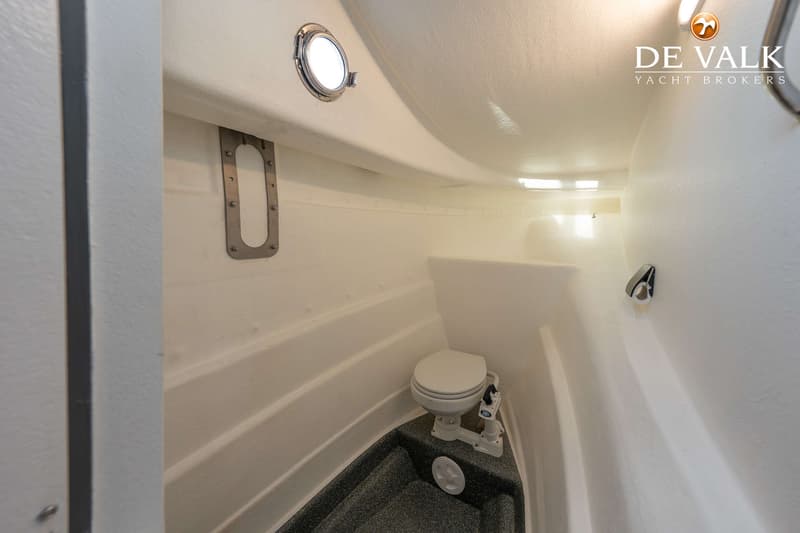 Een witte bootkajuit met een toilet, een patrijspoort en een grijs gespikkeld vloeroppervlak.