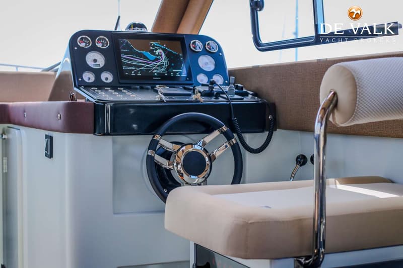 Een stuurstand van een boot met een stuur, navigatiescherm, meters en bedieningselementen, en een beige stoel.