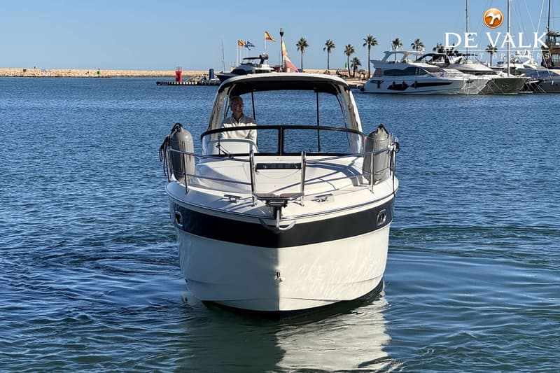 Een witte motorboot met een zwarte streep vaart op blauw water, met een man aan het roer en een haven op de achtergrond.