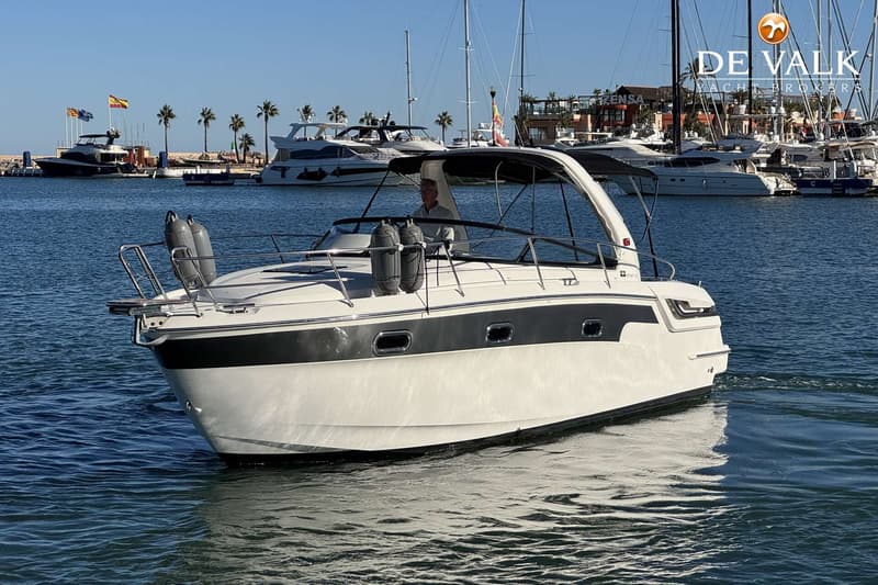 Een witte motorboot met een zwarte streep vaart op het water, met een man aan het roer en een haven op de achtergrond.