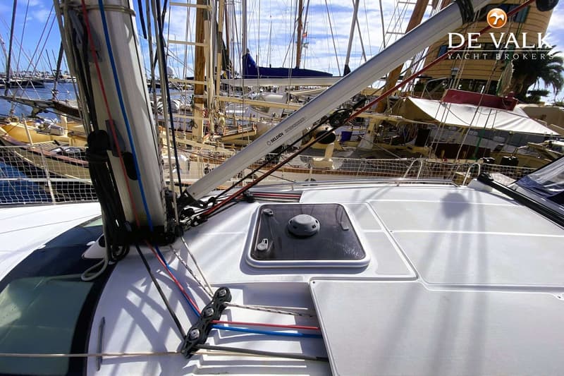De foto toont het dek van een zeilboot met een mast, giek, lijnen en een luik, met andere boten in de achtergrond.
